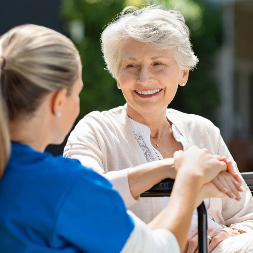 Pflegekraft spricht lächelnd mit einer älteren Frau im Rollstuhl im Freien